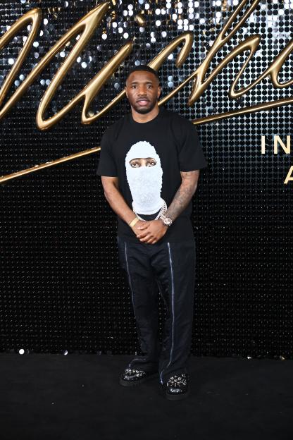 Konan posing on a black carpet in a black T-shirt and black pants with silver zipper details, in front of a black sequined backdrop with a gold "Michael" logo.