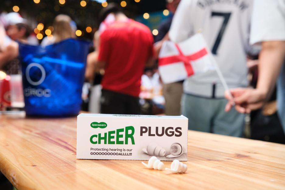 A box of Specsavers "Cheer Plugs" earplugs on a wooden table, with blurry background showing a person holding an England flag and other spectators.