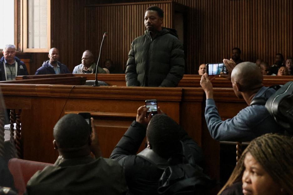 Ndodana Mkhanyisi Tshuma, wanted for murder, stands in a courtroom while spectators record him on their phones.