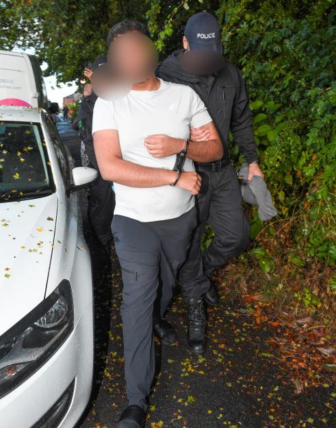 A handcuffed man in a white T-shirt being escorted by police officers.