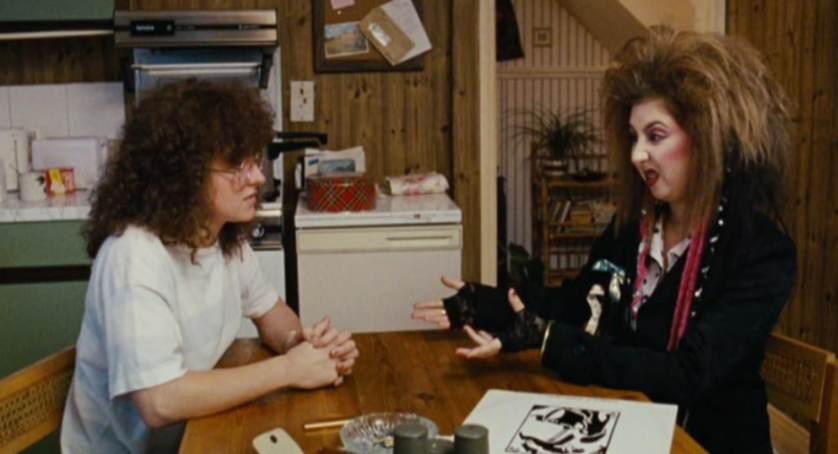 Two women sitting at a wooden table, one with curly hair and glasses, and the other with big hair and dark makeup, gesturing with her hands.