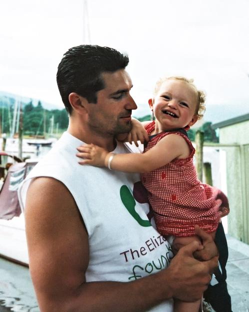 A man holding a young child with curly hair in a red checkered dress.