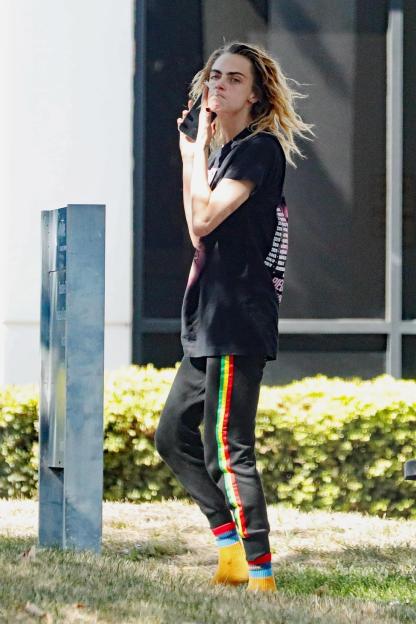 Cara Delevingne in a black t-shirt and track pants with colored stripes, holding a phone to her face, at Van Nuys Airport.