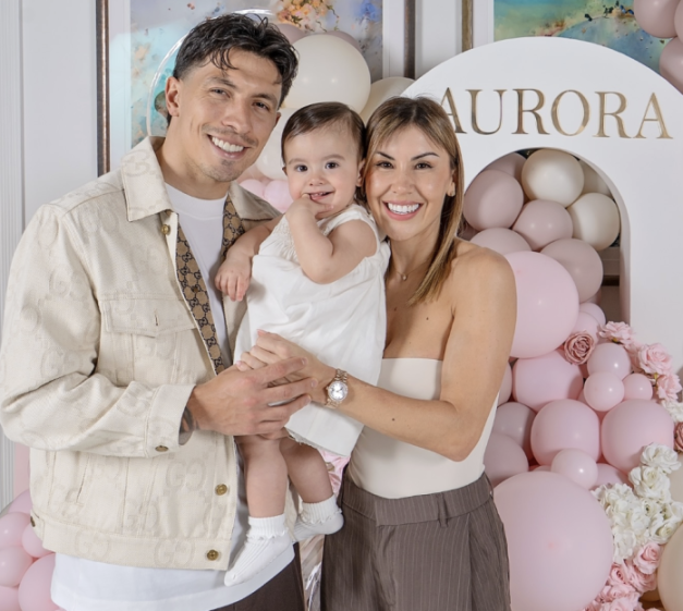 A man, woman, and baby stand in front of a balloon arch with the name "Aurora" on it.