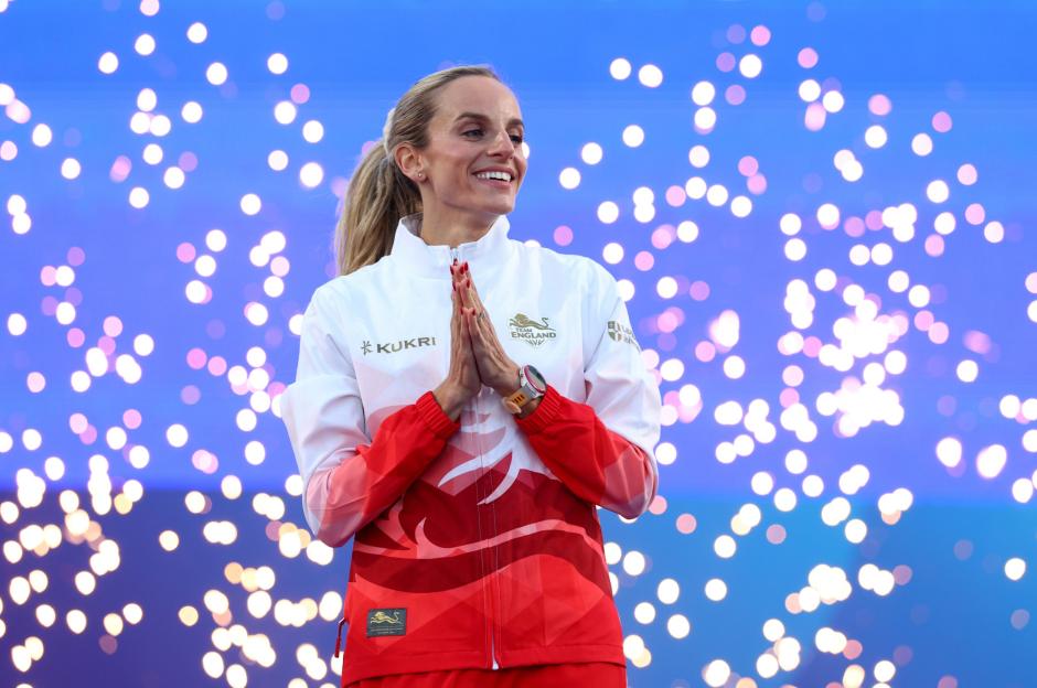 A woman in a white and red athletic jacket smiles with her hands clasped.