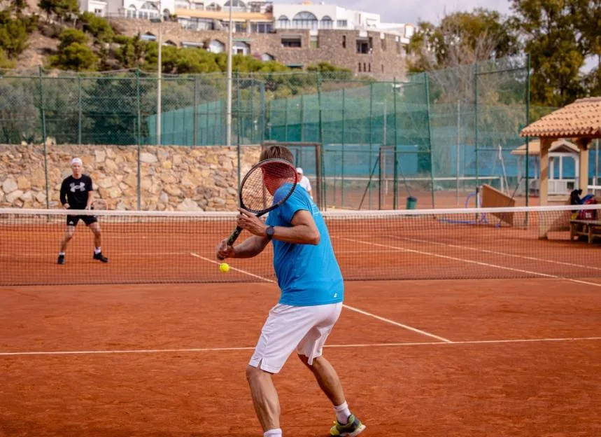 A man in a blue shirt prepares to hit a tennis ball on a clay court with another player waiting across the net.