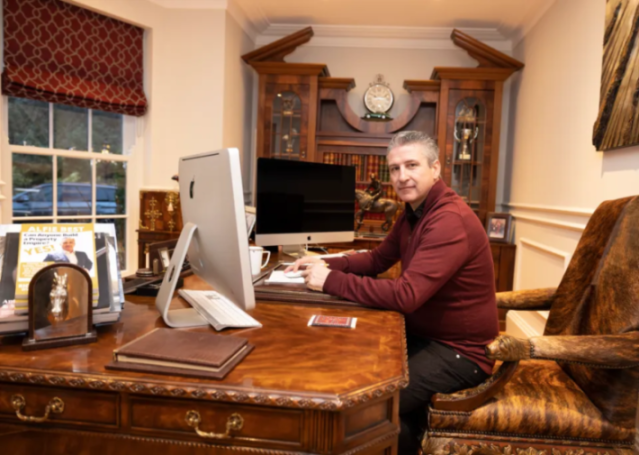 A man in a red sweater sitting at a large wooden desk with two iMacs, a horse statue, and a book titled "Can Anyone Build a Property Empire? YES!"