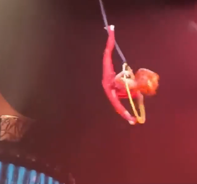 An aerialist in a red suit hangs upside down from a trapeze.
