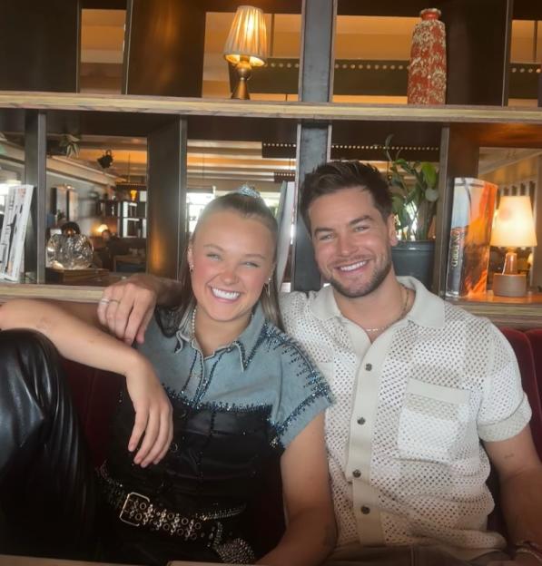 Jojo Siwa and Chris Hughes smiling and sitting on a red couch.