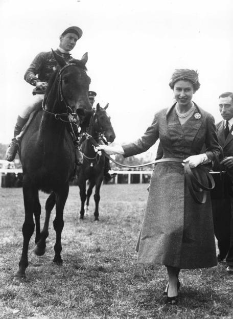 Queen Elizabeth II, wearing a formal dress and pearl necklace, leads her horse "Carrozza" with jockey Lester Piggott on its back, after winning the Oaks at Epsom.