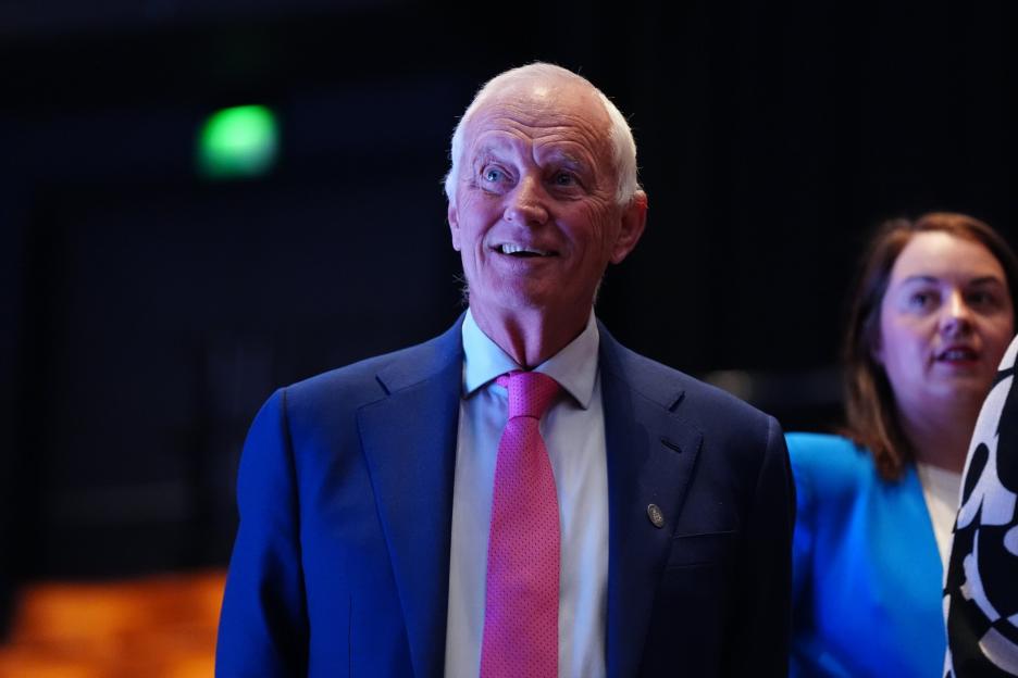 An older man with white hair, wearing a blue suit and a pink tie, smiling and looking up.