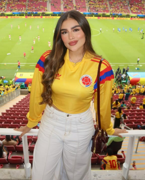 A woman wearing a yellow Colombian soccer jersey and white pants stands in a stadium overlooking a soccer match.