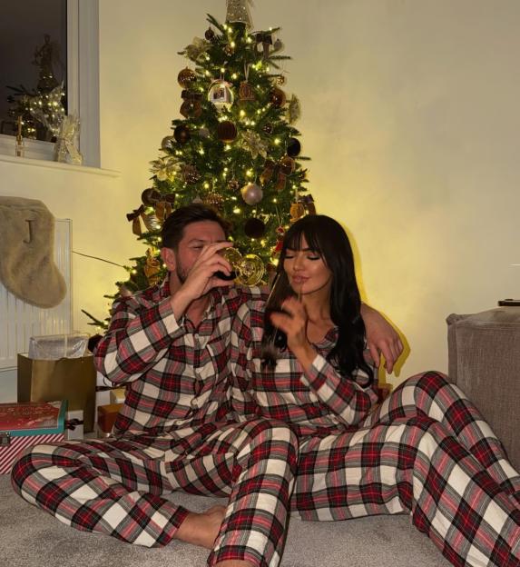 A couple in matching plaid pajamas sit on the floor in front of a Christmas tree.
