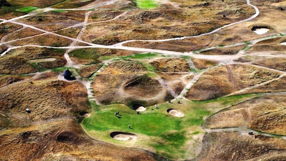An aerial view of a golf course with brown, dried-out fairways and green greens, with several golfers present.