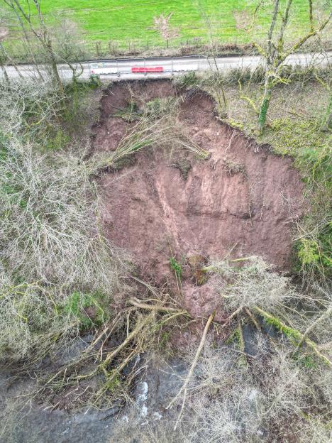 Aerial view of a landslide, showing a section of road collapsed into a ravine, with fallen trees and a river below.