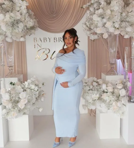 A pregnant woman in a light blue dress standing in front of a backdrop with white floral arrangements and a sign reading "BABY BROON".