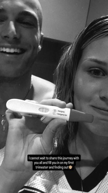 A black and white image of a smiling woman holding up a positive pregnancy test, with a man smiling in the background.