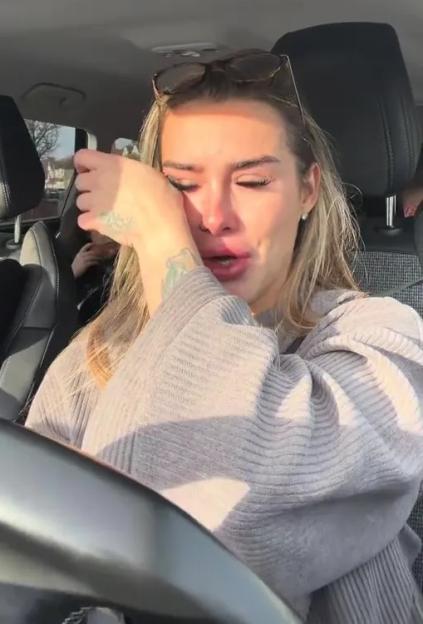 A woman with sunglasses on her head sits in a car, crying and wiping her eyes.