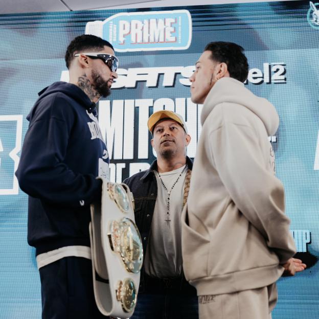 Two boxers and a man with a gold-colored belt in between them on a stage with a blue Prime logo in the background.