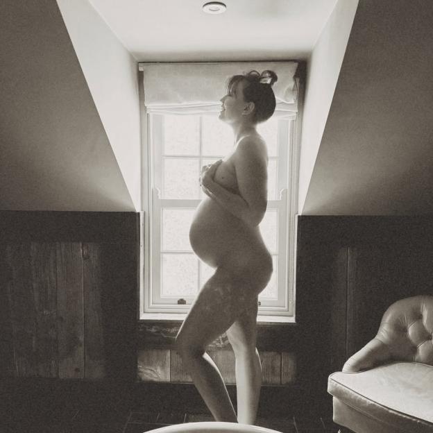 A pregnant woman stands naked in profile, holding her hands over her breasts, silhouetted against a sunlit window.