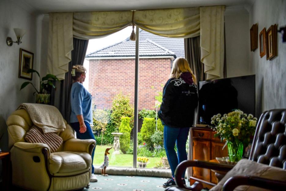 Two women look out of a window at a brick wall and green garden, with furnishings inside.