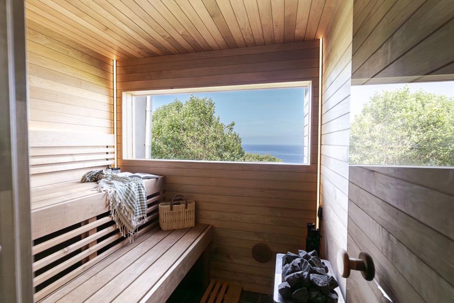 A sauna with wooden walls and benches, a window overlooking the ocean and trees, and a basket on one of the benches.