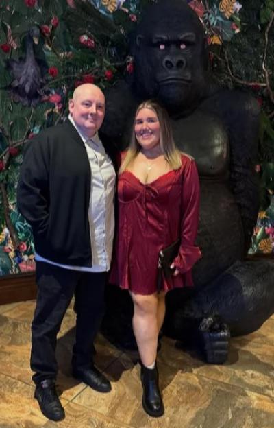 A man and woman standing next to a large gorilla statue in front of a floral wall.