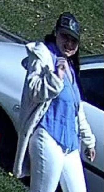 CCTV image of a woman in a blue shirt, white jeans, gray jacket, and baseball cap walking past a car.