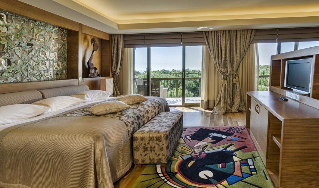 A spacious bedroom at Gloria Golf Resort with a large bed, decorative wall panels, a TV, and a balcony overlooking lush greenery.
