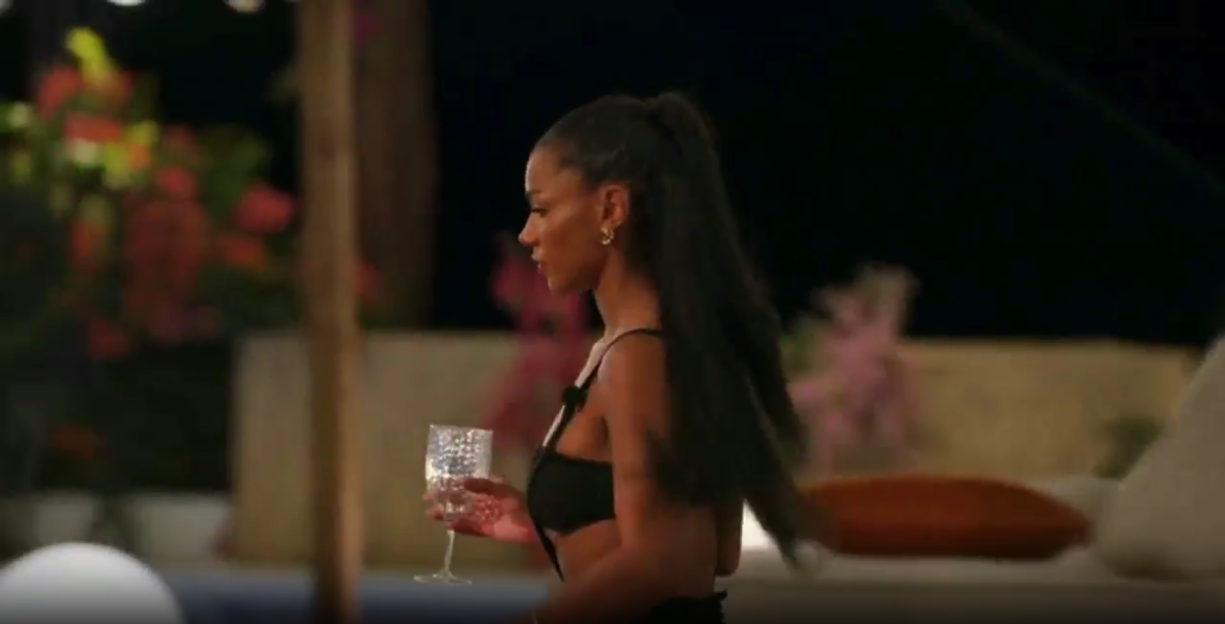 A woman with long dark hair in a black top holding a glass, walking at night.