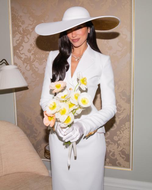 Dua Lipa in a white suit, white hat, and white gloves, holding a bouquet of light-colored flowers.