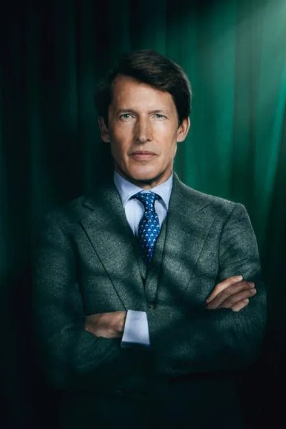James Blunt wearing a tweed suit and patterned tie with arms crossed against a green curtain background.