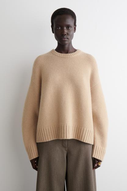 A dark-skinned model wearing a light beige crewneck sweater and dark gray pants stands in front of a white wall.
