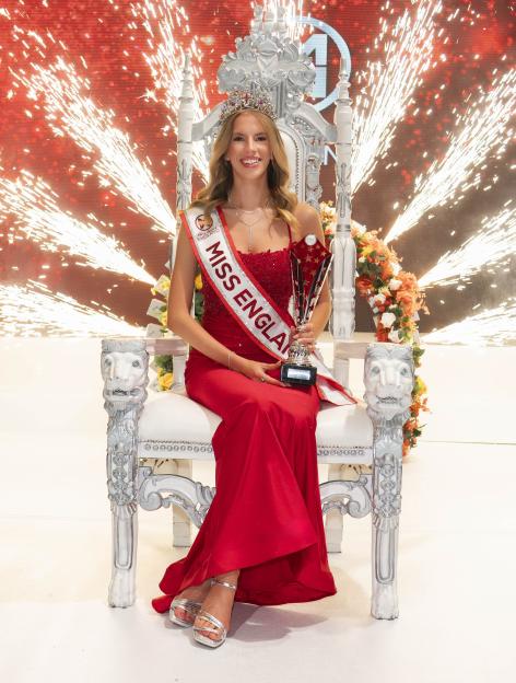 Grace Richardson, winner of Miss England 2025, sitting on a throne wearing a red dress, tiara, and sash, holding a trophy.