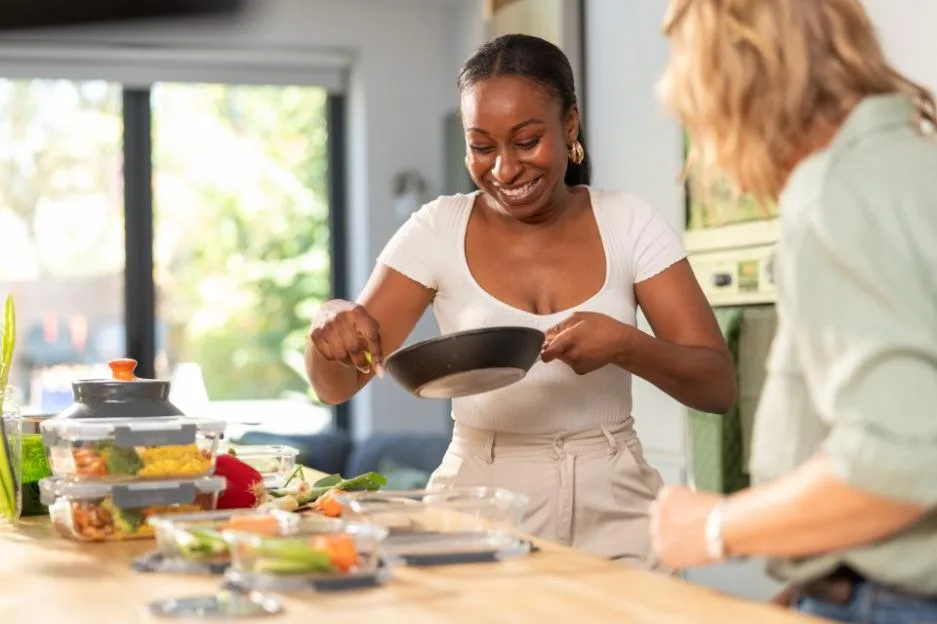 Joanna Page and another woman cooking in a kitchen, smiling and using a frying pan.