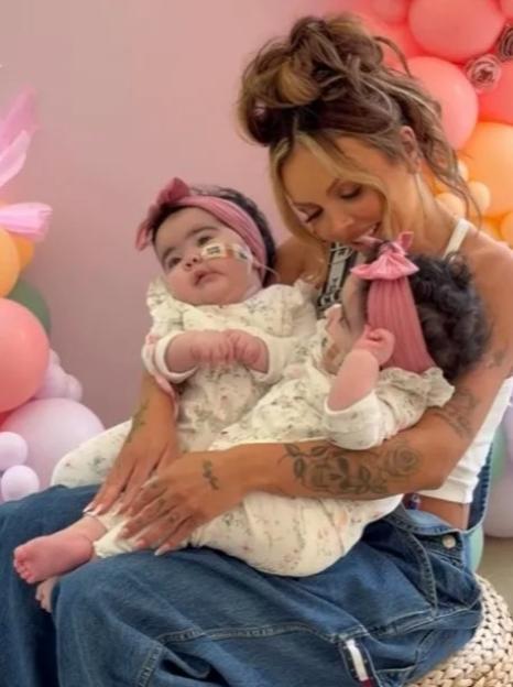Jesy Nelson holding her twin babies who are wearing matching floral outfits and pink headbands.