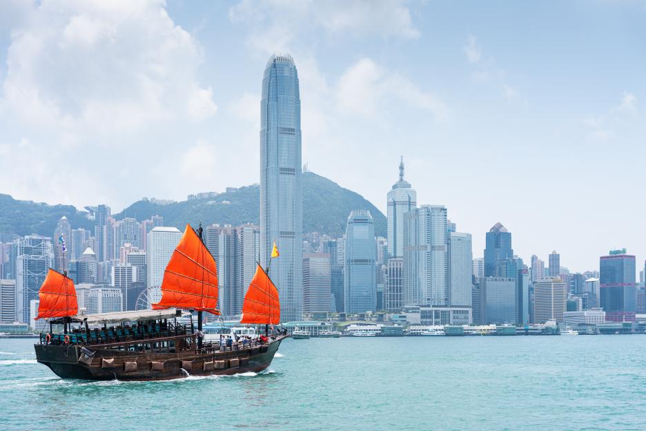An antique sailboat with red sails on Victoria Harbour in Hong Kong, with the city's skyline in the background.