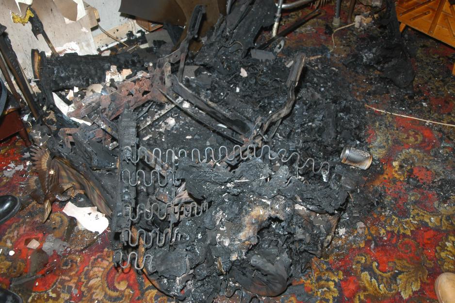 The charred remains of a chair, with springs visible, after a house fire on a floral patterned rug.