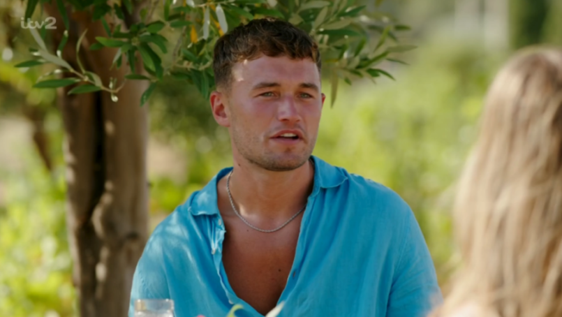 A man in a blue shirt with short brown hair, wearing a silver chain, looks off to the side.