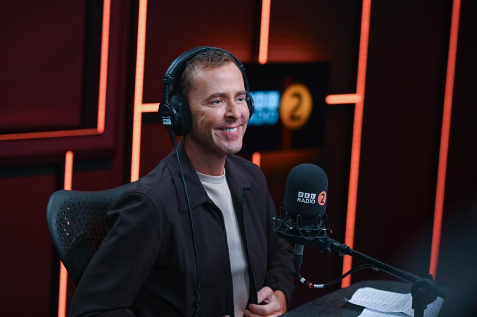 Scott Mills smiling while wearing headphones and sitting in front of a BBC Radio 2 microphone.