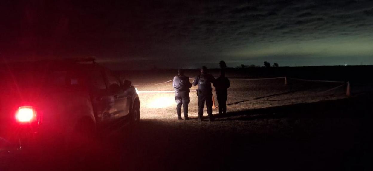 Three individuals standing in a dark field at night, illuminated by a car's headlights and taillights.