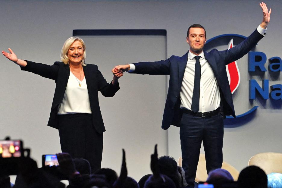 French far-right politicians Marine Le Pen and Jordan Bardella holding hands and waving to a crowd.