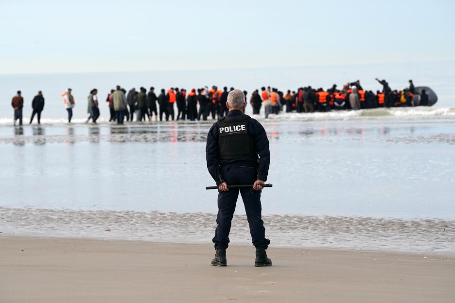 A police officer on a beach watches migrants in the water boarding a small boat.
