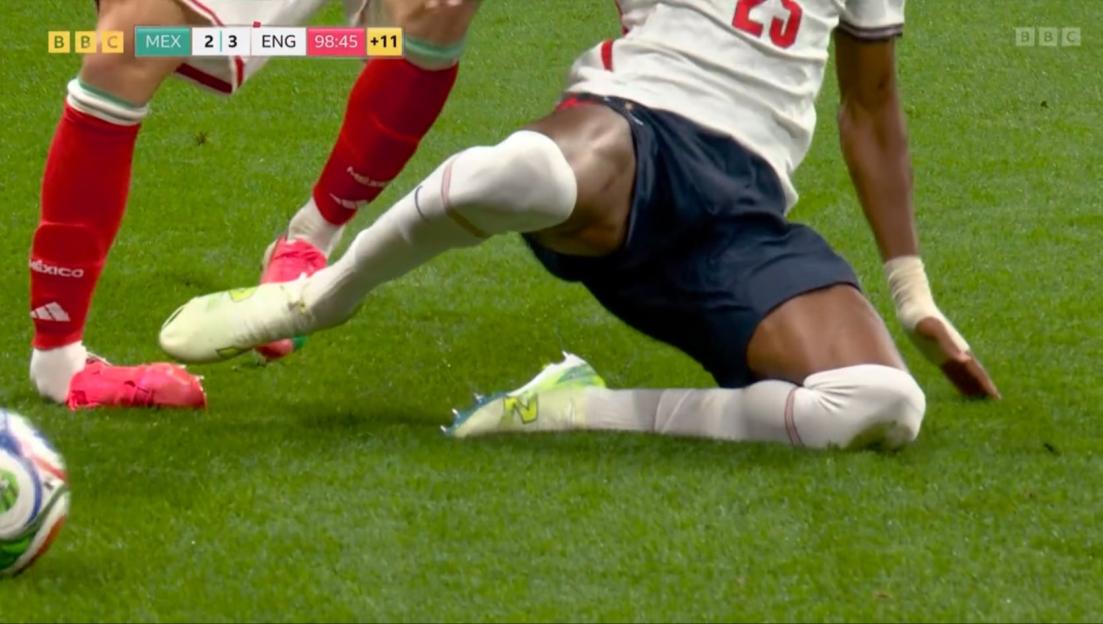 A soccer player in a white and blue uniform slides on the grass with another player in red and green nearby.