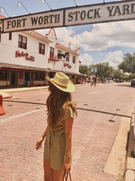A person with long brown hair and a straw cowboy hat stands on a brick road under a sign reading "Fort Worth Stock Yard."