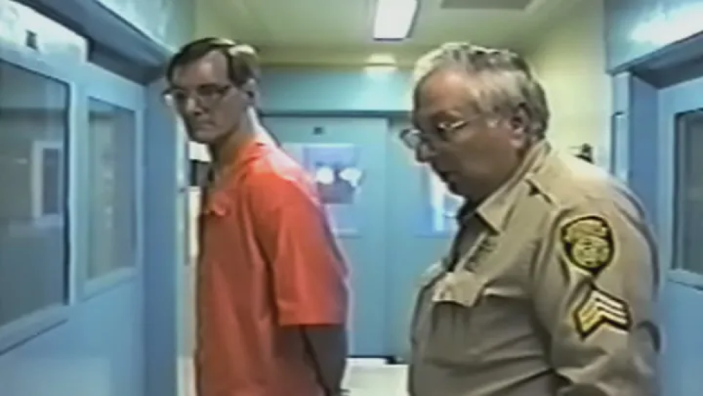 Serial killer Doug Gretzler in an orange jumpsuit with an officer in a prison hallway.