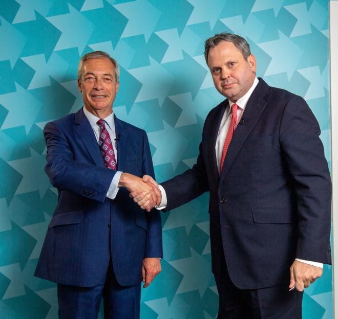Nigel Farage and Harry Cole shake hands, both men wearing blue suits and ties, in front of a blue background with a star pattern.