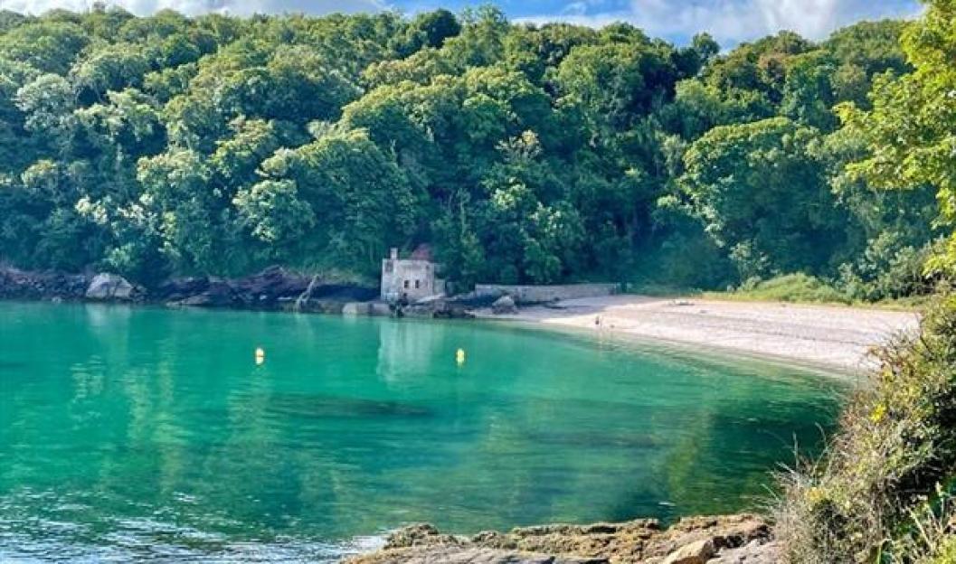 Elberry Cove with turquoise water, a pebble beach, and a small stone building nestled at the base of a forested hill.
