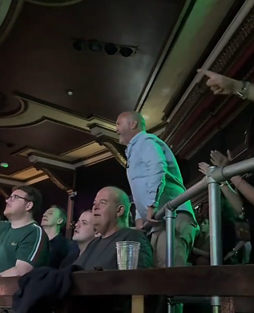 Man stands up and cheers in a balcony seat at a concert.