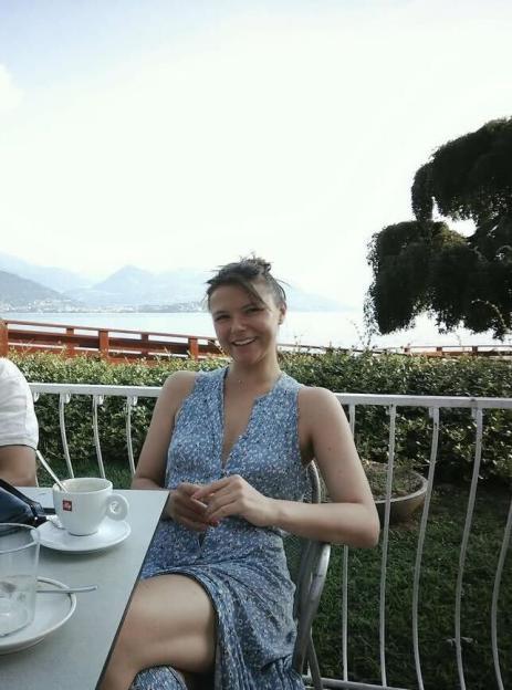 A young woman in a blue floral dress smiles at a cafe table overlooking a lake and mountains.
